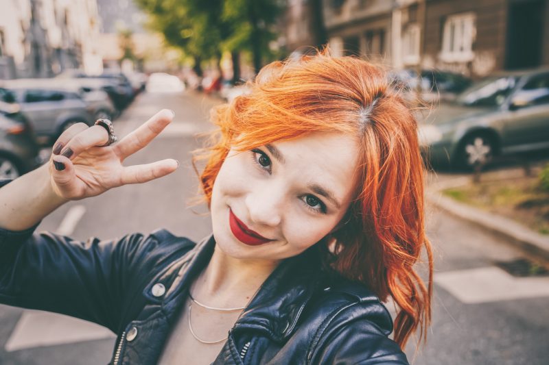 Selfie of a woman with copper hair smiling and giving the peace sign on the street outside. Find your perfect red hair color viviscal hair blog