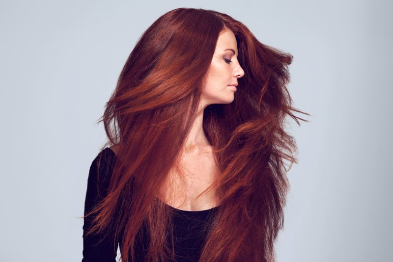 Woman shaking her long dark red auburn hair against a gray background. find your perfect red hair color viviscal hair blog