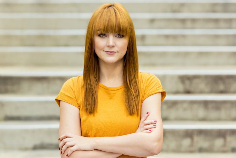 redhead woman with blunt bangs and long hair stands outside in an orange shirt.