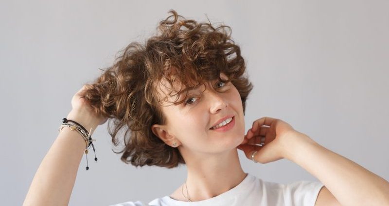 woman tosses her head and short, curly, brown hair and bangs to the right.